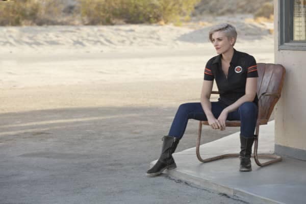A women with short blond hair sitting in a chair wearing a black V-neck polo from the Harley-Davidson Genuine MotorClothes collection.