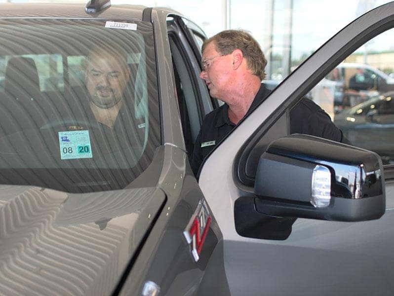 An exterior shot of a customer sitting behind the wheel of a car while a Sales Representative helps out