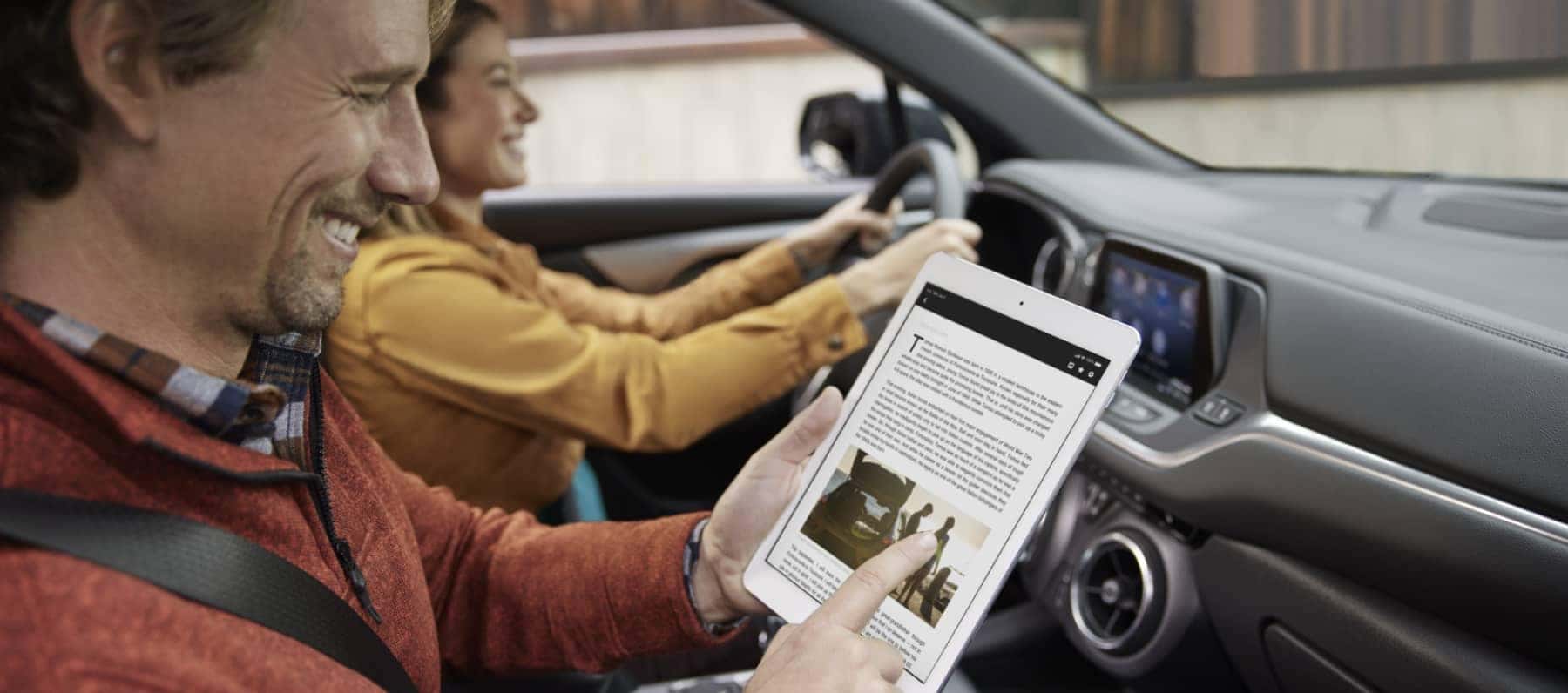 man is smiling while looking at a tablet in the passenger seat of car