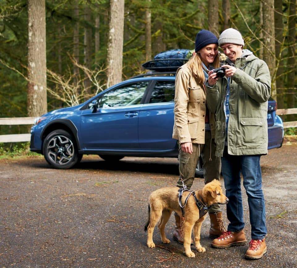 Couple and dog in nature looking at a pictures on a camera with a parked blue Subaru behind them