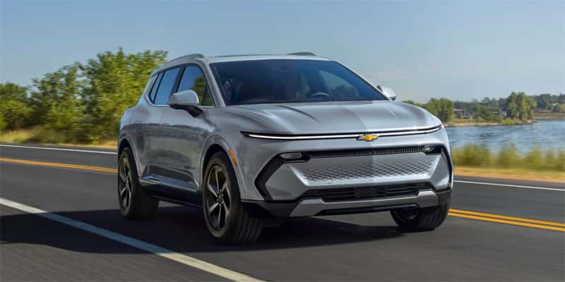 An all-new silver 2025 Chevy Equinox EV driving along a waterside highway. - Toliver Brothers Chevrolet Ballinger, TX