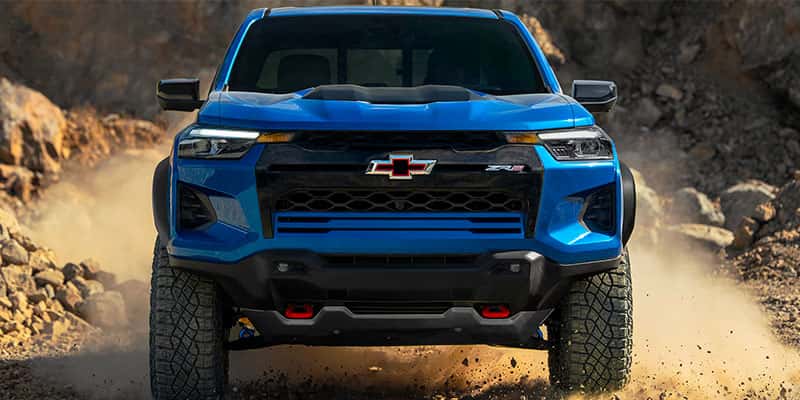 An all-new blue 2025 Chevy Colorado from the front view, driving across a sand and rock terrain. - Toliver Brothers Chevrolet