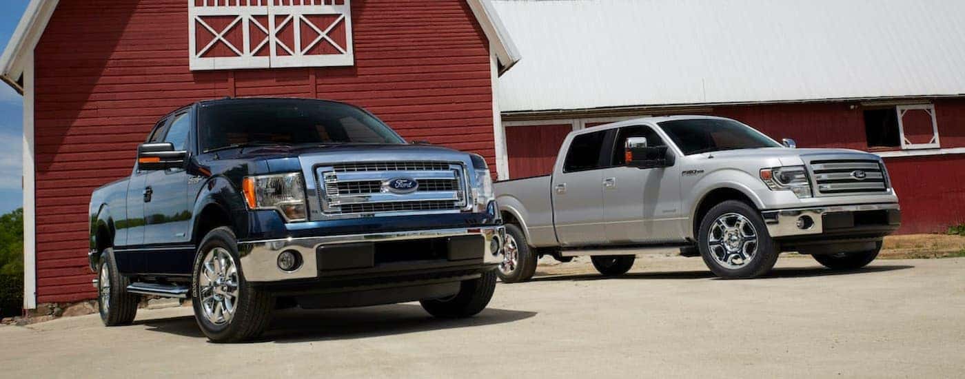 A black 2014 Ford F-150 XLT and silver Lariat are shown parked next to each other in front of a red barn.