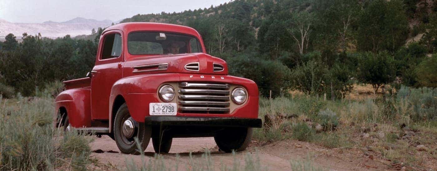 A red 1948 Ford F-1 is parked on a dirt path near a wooded hill.