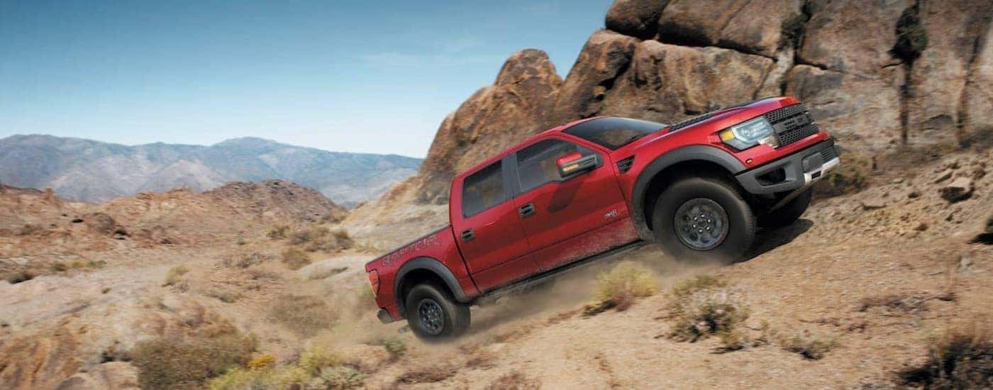 A red 2014 Ford Raptor SVT is shown driving up a dusty hill past a rock face.