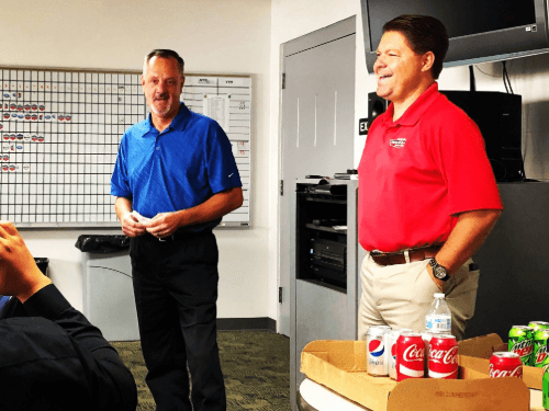 Men standing in a room with a box of soda and a person in a blue shirt