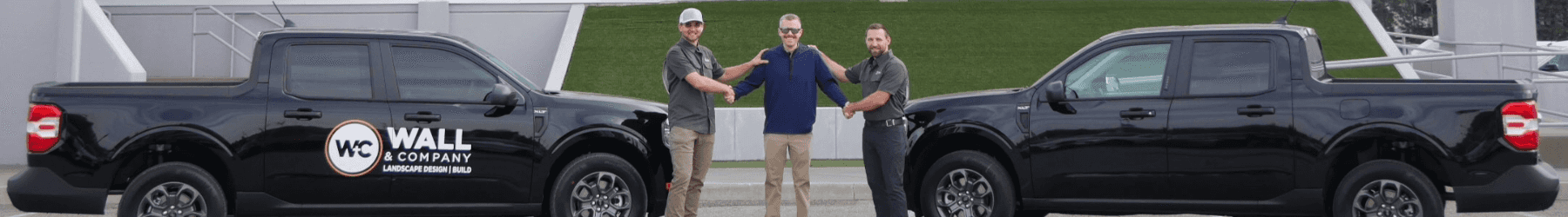 Three men standing in front of car at a dealership with two black trucks.