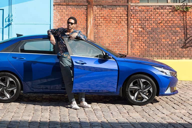 A man standing in the open passenger door of a 2021 Intense Blue Elantra parked in front of a brick building